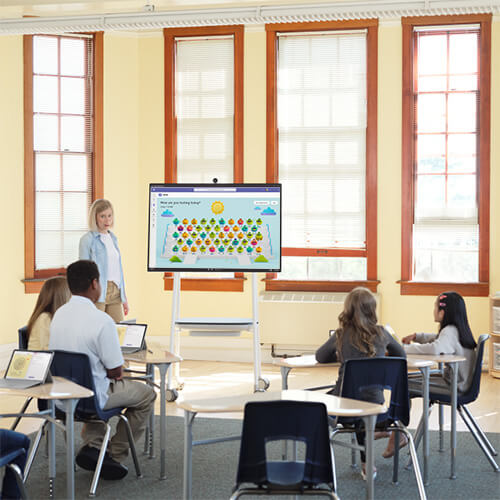 Several school age children are sitting at desks with laptops open on their desks looking at the teacher and Hub with Reflect at the front of the classroom